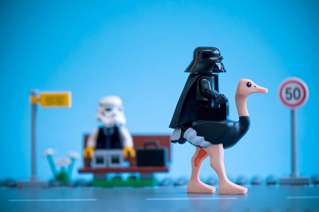 A Lego Darth Vader riding an ostrich on a road wiht a blurry stormtrooper masked man sitting on a bench